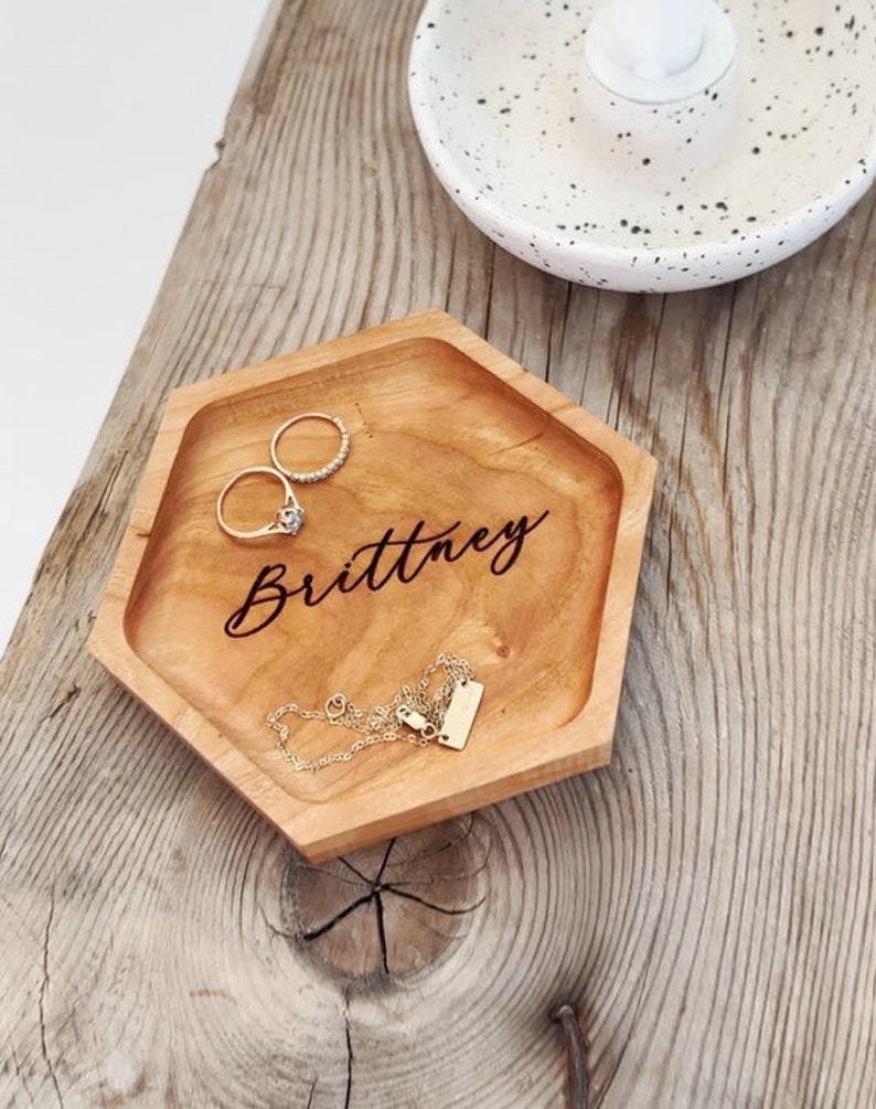 May include: A hexagonal wooden tray with the name "Brittney" engraved on it. The tray is filled with jewelry, including two rings and a necklace.