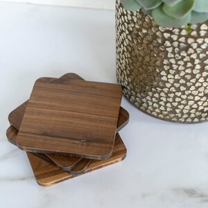 May include: Four square wooden coasters stacked on top of each other. The coasters are made of dark brown wood with a natural grain pattern.