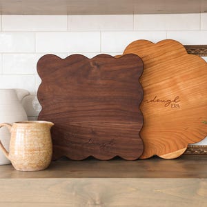 May include: Two wooden cutting boards with scalloped edges. The larger board is made of cherry wood and has the text "Wanderlust Era" engraved on it. The smaller board is made of walnut wood.