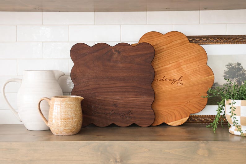 May include: Two wooden cutting boards with scalloped edges. The larger board is made of cherry wood and has the text "Wanderlust Era" engraved on it. The smaller board is made of walnut wood.