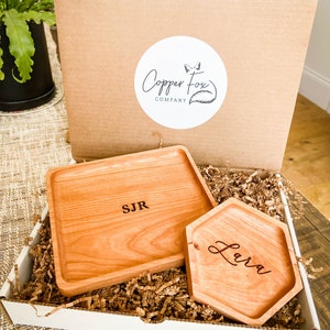 May include: Two wooden trays with personalized engravings. The larger tray is rectangular and engraved with "SJR". The smaller tray is hexagonal and engraved with "Lara". Both trays are in a white box with brown packing paper.