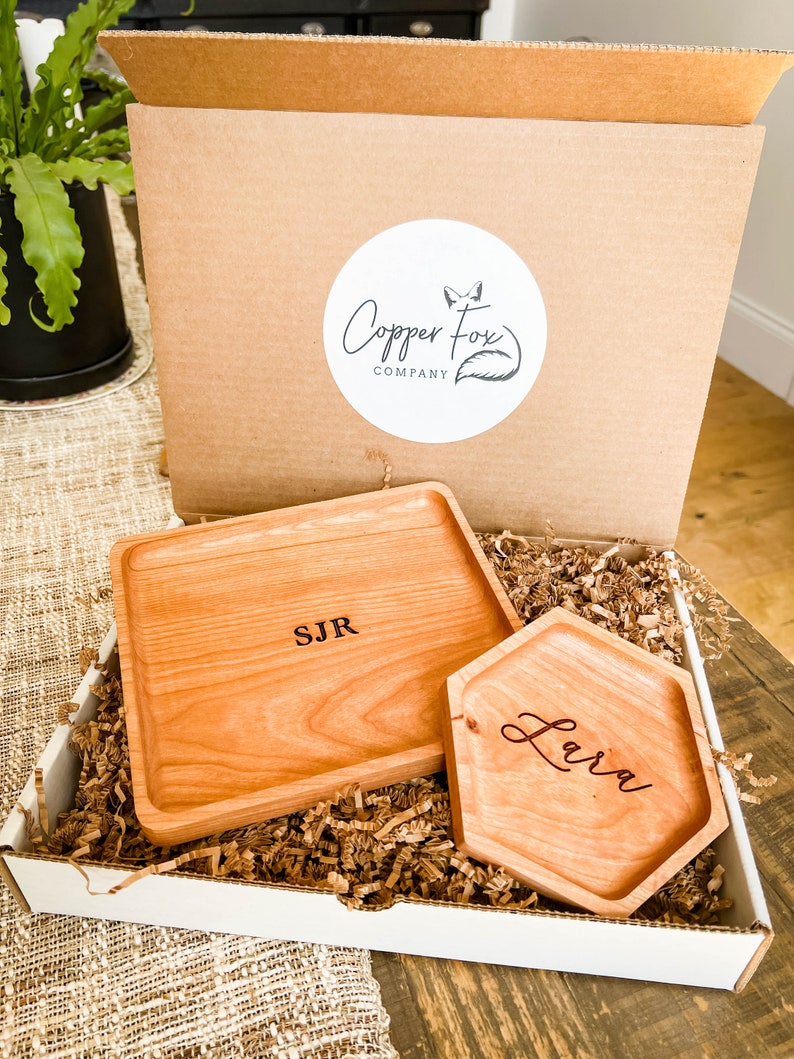 May include: Two wooden trays with personalized engravings. The larger tray is rectangular and engraved with "SJR". The smaller tray is hexagonal and engraved with "Lara". Both trays are in a white box with brown packing paper.