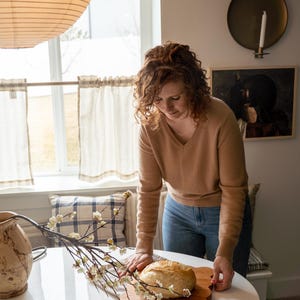 May include: A woman in a brown sweater and blue jeans places a loaf of sourdough bread on a wooden cutting board. The bread is on a white table with a sprig of white blossoms.