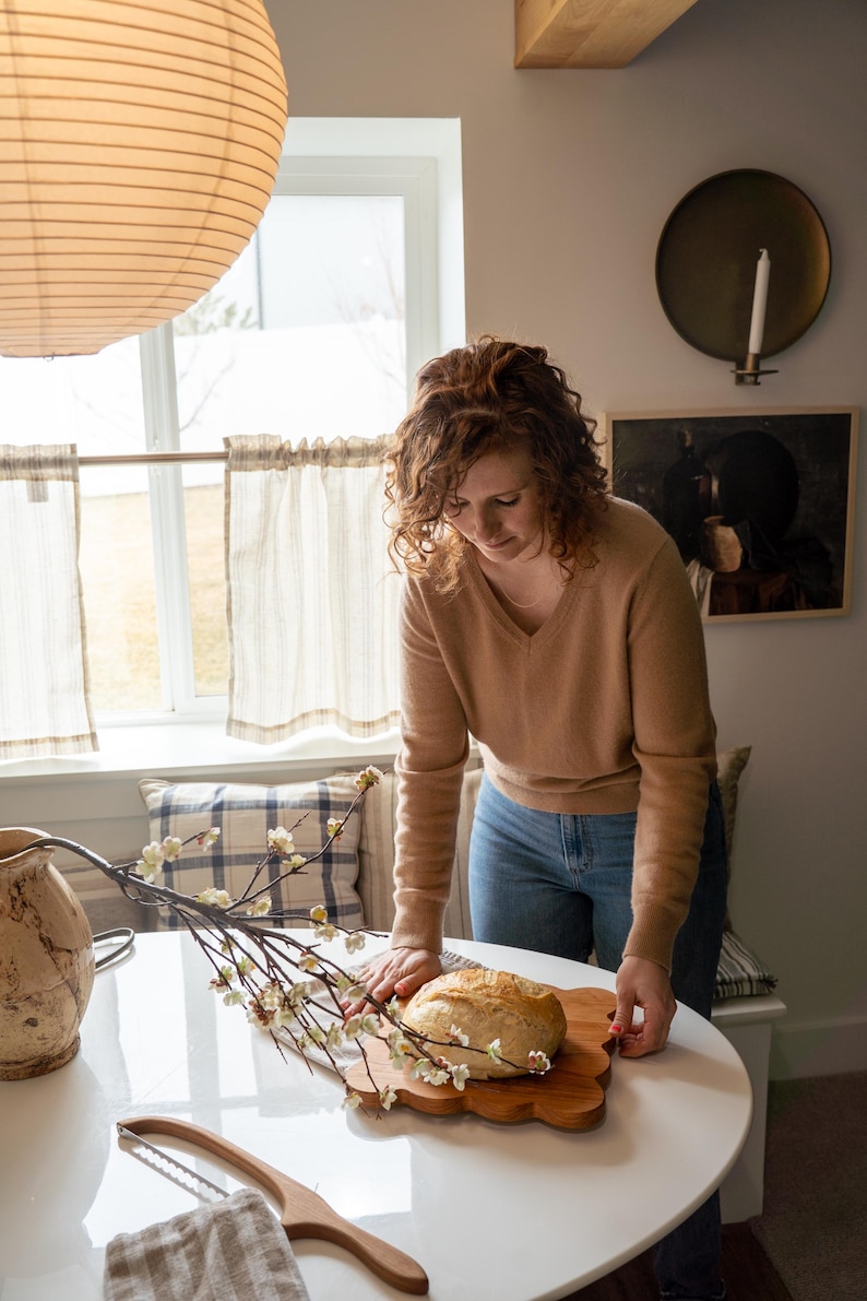 May include: A woman in a brown sweater and blue jeans places a loaf of sourdough bread on a wooden cutting board. The bread is on a white table with a sprig of white blossoms.