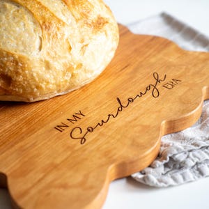May include: A wooden cutting board with a scalloped edge and the text "In My Sourdough Era" engraved on the surface. A loaf of sourdough bread is sitting on the board.