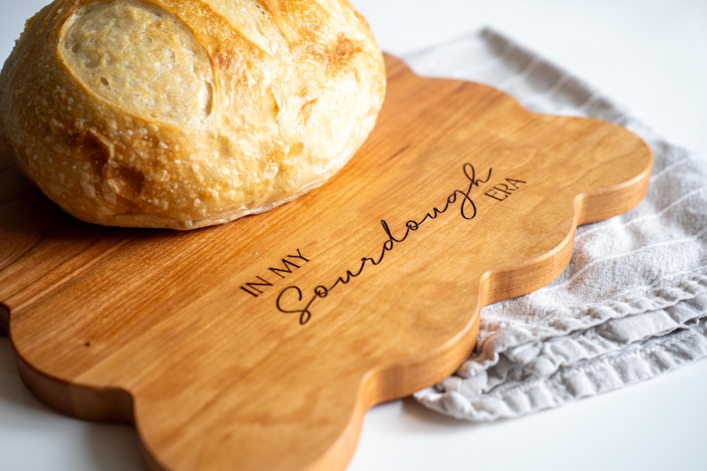 May include: A wooden cutting board with a scalloped edge and the text "In My Sourdough Era" engraved on the surface. A loaf of sourdough bread is sitting on the board.