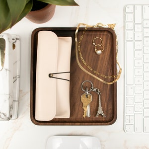 May include: A wooden tray with a light pink leather pouch, a gold chain necklace, two gold rings, a pearl, a set of keys with a silver Eiffel Tower keychain, and a white computer keyboard.