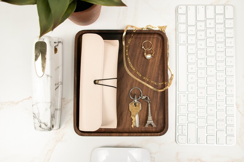 May include: A wooden tray with a light pink leather pouch, a gold chain necklace, two gold rings, a pearl, a set of keys with a silver Eiffel Tower keychain, and a white computer keyboard.