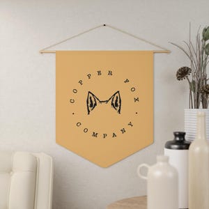 Op de afbeelding: Een beige stoffen banner met een zwart silhouet van een vossenkop en de tekst "Copper Fox Company" in een cirkel rond de vossenkop.