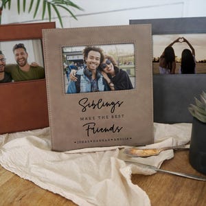 May include: Three rectangular photo frames in tan, brown, and gray with a smooth finish. The center frame features the text "Siblings MAKE THE BEST Friends" followed by the names "Jonas • Hannah • Amelia" printed in black ink below a photo.