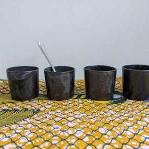 May include: Four small, cylindrical black cups with a textured surface are arranged on a patterned yellow and blue cloth. One cup contains a silver spoon. The cups are likely for drinking.