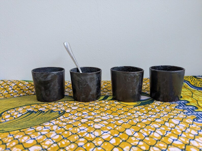 May include: Four small, cylindrical black cups with a textured surface are arranged on a patterned yellow and blue cloth. One cup contains a silver spoon. The cups are likely for drinking.