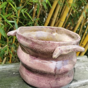 Puede incluir: Una maceta de cerámica con un diseño texturizado y en espiral en tonos marrones y morados. La maceta tiene dos pequeñas asas a los lados.