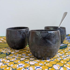 May include: Three dark, speckled ceramic cups with rounded bodies. One cup holds a silver spoon. The cups are on a yellow and blue patterned fabric, against a white background. The cups appear handmade.
