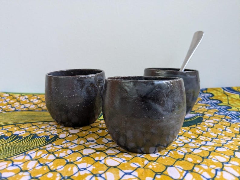 May include: Three dark, speckled ceramic cups with rounded bodies. One cup holds a silver spoon. The cups are on a yellow and blue patterned fabric, against a white background. The cups appear handmade.