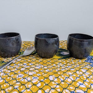 May include: Three dark gray ceramic cups with a textured surface are arranged on a yellow and blue patterned cloth. A silver spoon rests between the cups. The cups have a unique, handcrafted appearance.