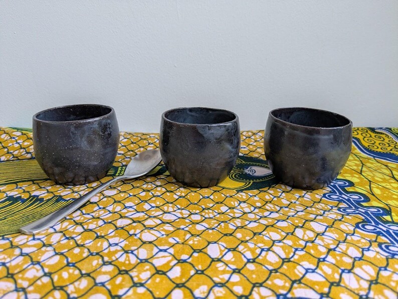 May include: Three dark gray ceramic cups with a textured surface are arranged on a yellow and blue patterned cloth. A silver spoon rests between the cups. The cups have a unique, handcrafted appearance.