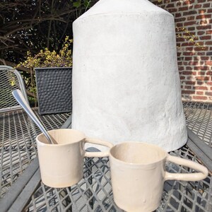 May include: Two beige ceramic mugs with handles on a metal table. A silver spoon is on the table. A large white ceramic vase is in the background.