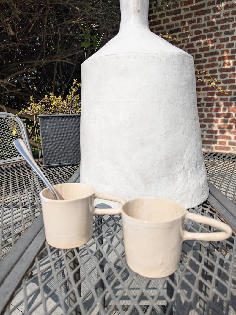 May include: Two beige ceramic mugs with handles on a metal table. A silver spoon is on the table. A large white ceramic vase is in the background.