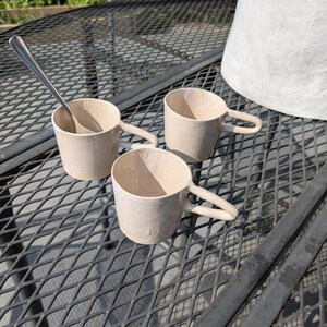 May include: Three white ceramic mugs with rounded handles on a black metal table. A silver spoon is resting in one of the mugs.