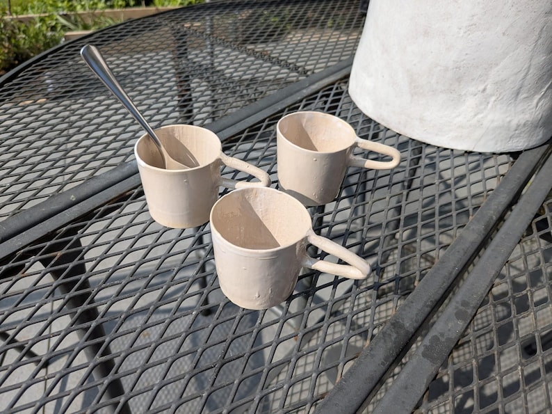 May include: Three white ceramic mugs with rounded handles on a black metal table. A silver spoon is resting in one of the mugs.