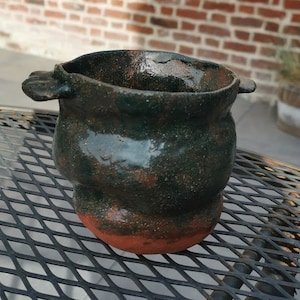 Może przedstawiać: Mały, zielono-brązowy ceramiczny garnek z dwoma uchwytami. Garnek ma fakturowaną powierzchnię i stoi na czarnym metalowym stole.