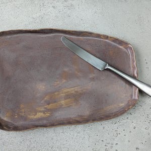 K&ouml;nnte beinhalten: Ein braunes Keramik-Serviertablett mit unregelm&auml;&szlig;iger Form und leicht strukturierter Oberfl&auml;che. Ein silbernes Messer liegt auf dem Tablett.