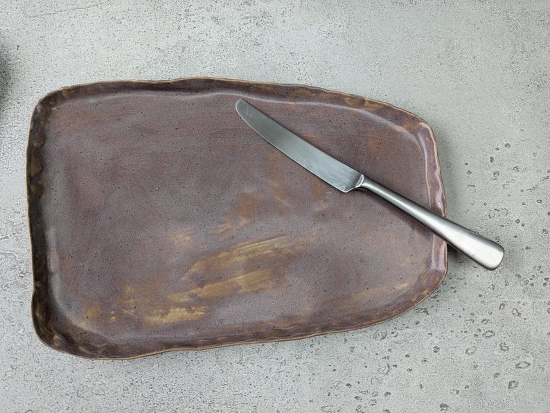 K&ouml;nnte beinhalten: Ein braunes Keramik-Serviertablett mit unregelm&auml;&szlig;iger Form und leicht strukturierter Oberfl&auml;che. Ein silbernes Messer liegt auf dem Tablett.