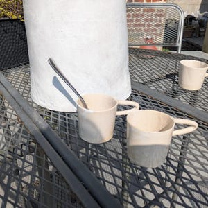 May include: Three ceramic mugs with a light beige glaze sit on a black metal table with a diamond pattern. The mugs have rounded handles and are slightly different sizes. A silver spoon is lying on the table.