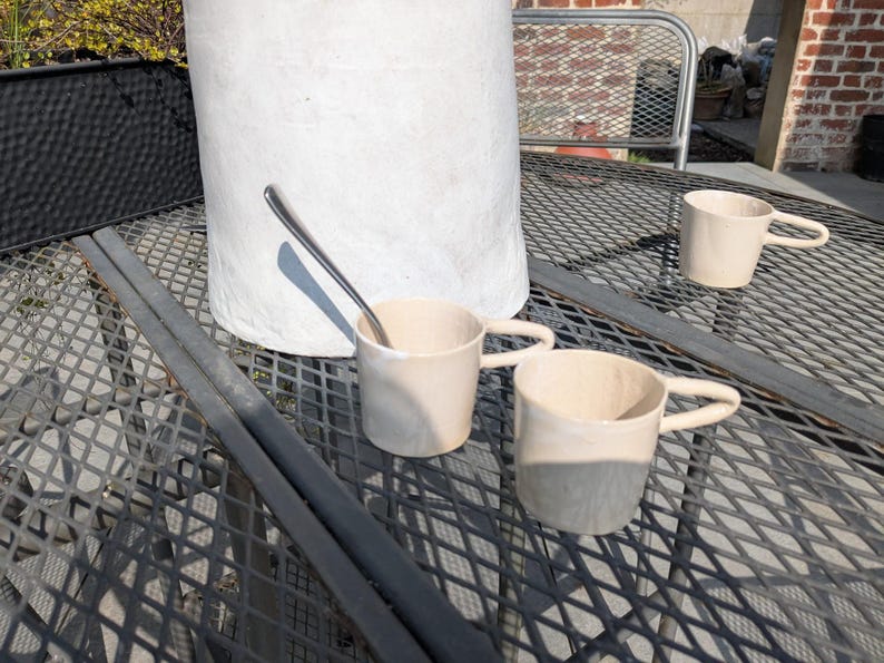 May include: Three ceramic mugs with a light beige glaze sit on a black metal table with a diamond pattern. The mugs have rounded handles and are slightly different sizes. A silver spoon is lying on the table.