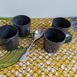 May include: Four small, dark gray metal cups and a silver spoon are arranged on a yellow and blue patterned cloth. The cups have a rustic, textured appearance, and the spoon is smooth and reflective.