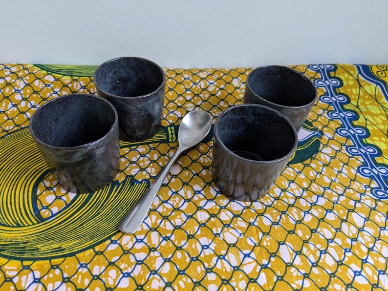 May include: Four small, dark gray metal cups and a silver spoon are arranged on a yellow and blue patterned cloth. The cups have a rustic, textured appearance, and the spoon is smooth and reflective.