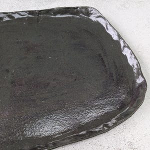 K&ouml;nnte beinhalten: Ein schwarzes Keramik-Serviertablett mit leicht unregelm&auml;&szlig;iger, abgerundeter Form. Das Tablett hat eine glatte, gl&auml;nzende Oberfl&auml;che.