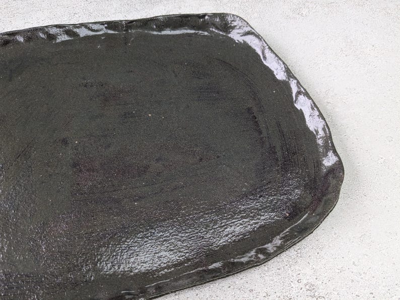 K&ouml;nnte beinhalten: Ein schwarzes Keramik-Serviertablett mit leicht unregelm&auml;&szlig;iger, abgerundeter Form. Das Tablett hat eine glatte, gl&auml;nzende Oberfl&auml;che.