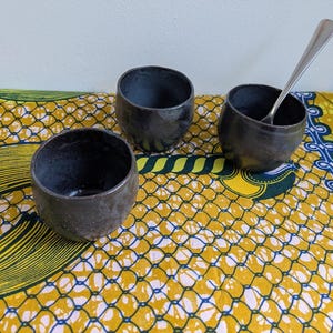 May include: Three dark gray ceramic bowls with a textured surface are arranged on a colorful patterned cloth. One bowl contains a silver spoon. The bowls are round with a slightly irregular shape, suggesting they are handmade.