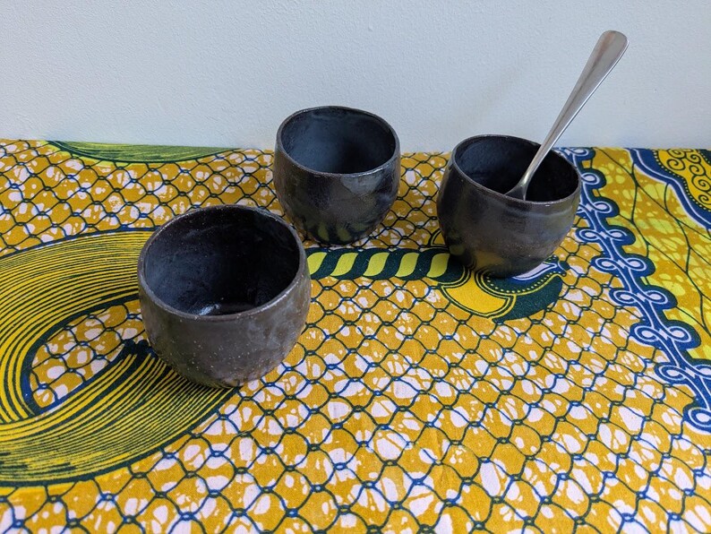 May include: Three dark gray ceramic bowls with a textured surface are arranged on a colorful patterned cloth. One bowl contains a silver spoon. The bowls are round with a slightly irregular shape, suggesting they are handmade.