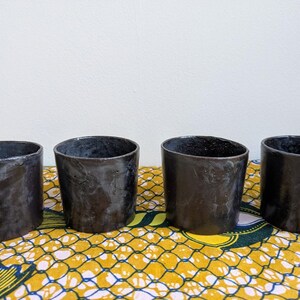 May include: Four handmade, dark charcoal-colored ceramic cups with a slightly irregular shape. The cups are arranged in a row on a patterned yellow and blue fabric, with a plain white background.