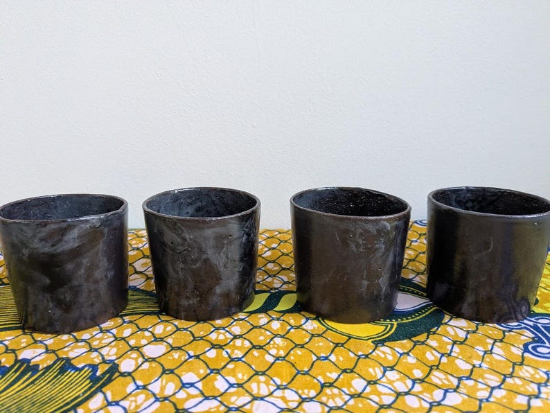 May include: Four handmade, dark charcoal-colored ceramic cups with a slightly irregular shape. The cups are arranged in a row on a patterned yellow and blue fabric, with a plain white background.