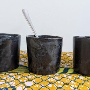 May include: Three handmade, dark charcoal-colored ceramic cups with a rough texture. One cup holds a silver spoon. The cups are cylindrical with slightly irregular rims. They sit on a yellow and green patterned fabric.