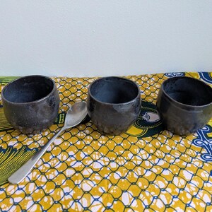 May include: Three dark gray ceramic bowls with a textured surface are arranged on a colorful yellow, white, and blue patterned cloth. A silver spoon rests between the bowls. The bowls have a rustic, handmade appearance.