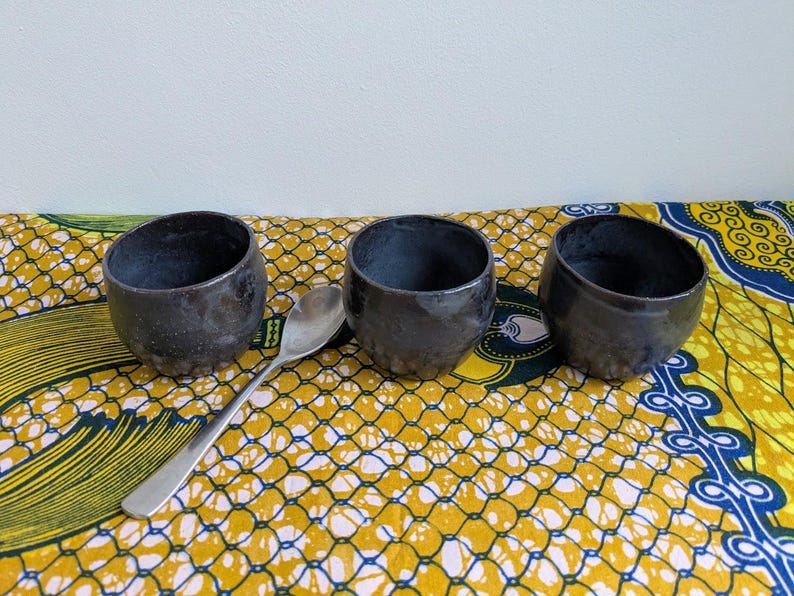 May include: Three dark gray ceramic bowls with a textured surface are arranged on a colorful yellow, white, and blue patterned cloth. A silver spoon rests between the bowls. The bowls have a rustic, handmade appearance.