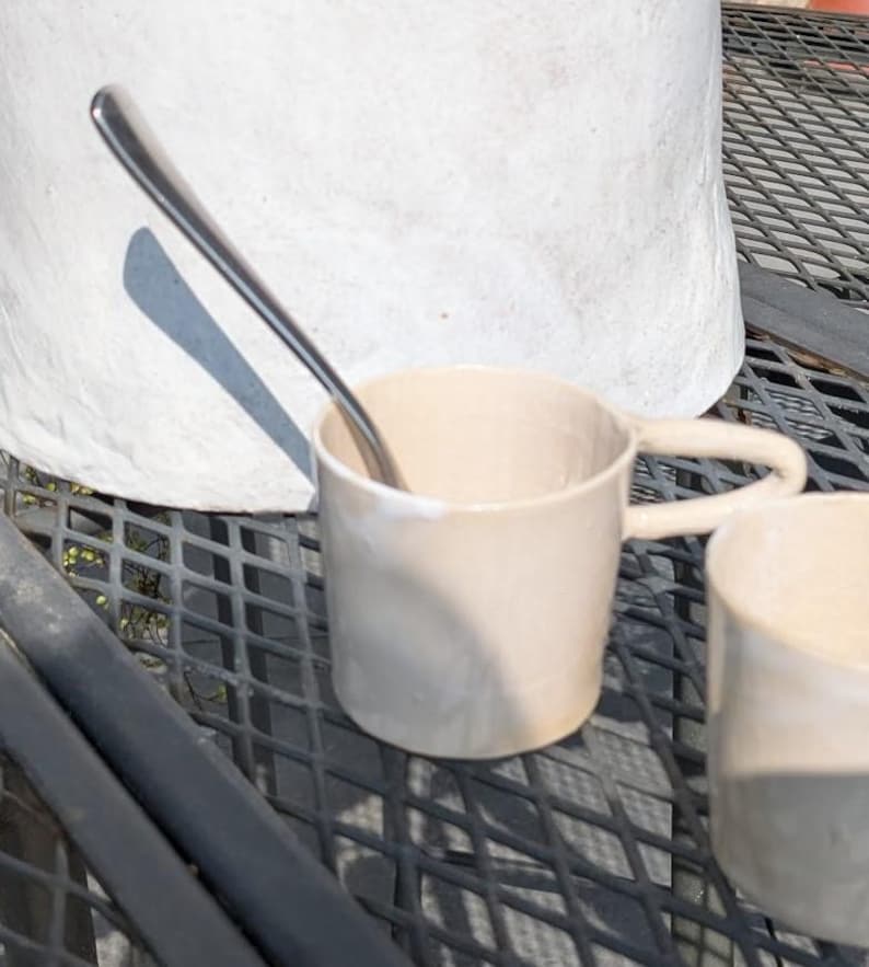 May include: Two beige ceramic mugs with a silver spoon resting on the rim of one mug. The mugs are on a black metal table.