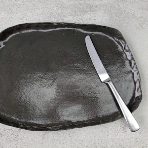 K&ouml;nnte beinhalten: Eine schwarze Keramik-Servierplatte mit leicht unregelm&auml;&szlig;iger Form und glatter, gl&auml;nzender Oberfl&auml;che. Ein silbernes Messer liegt auf der Platte.