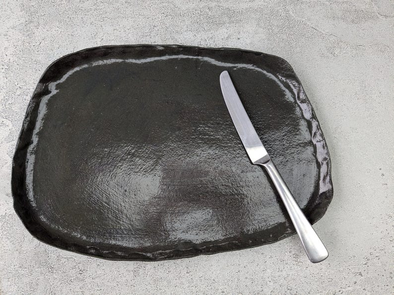 K&ouml;nnte beinhalten: Eine schwarze Keramik-Servierplatte mit leicht unregelm&auml;&szlig;iger Form und glatter, gl&auml;nzender Oberfl&auml;che. Ein silbernes Messer liegt auf der Platte.