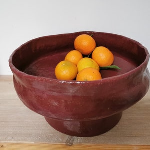 Peut inclure: Un bol en céramique rouge foncé avec un bord large et un piédestal. Le bol est rempli de petits agrumes orange vif.