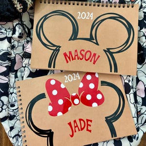 Könnte beinhalten: Zwei braune Spiralnotizbücher mit einem Mickey Mouse Ohren-Design und dem Jahr 2024. Ein Notizbuch hat den Namen "Mason" in Rot, das andere eine rote Schleife mit weißen Punkten und den Namen "Jade". Eine schwarze Pailletten-Schleife und ein Minnie Mouse Rucksack sind im Hintergrund.