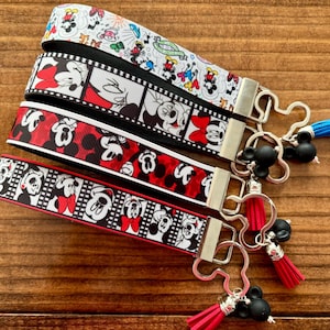 May include: Four key fobs with various Mickey Mouse and Minnie Mouse designs. The key fobs have silver-colored hardware and black Mickey Mouse-shaped charms with tassels. The ribbons feature red, black, and white colors.