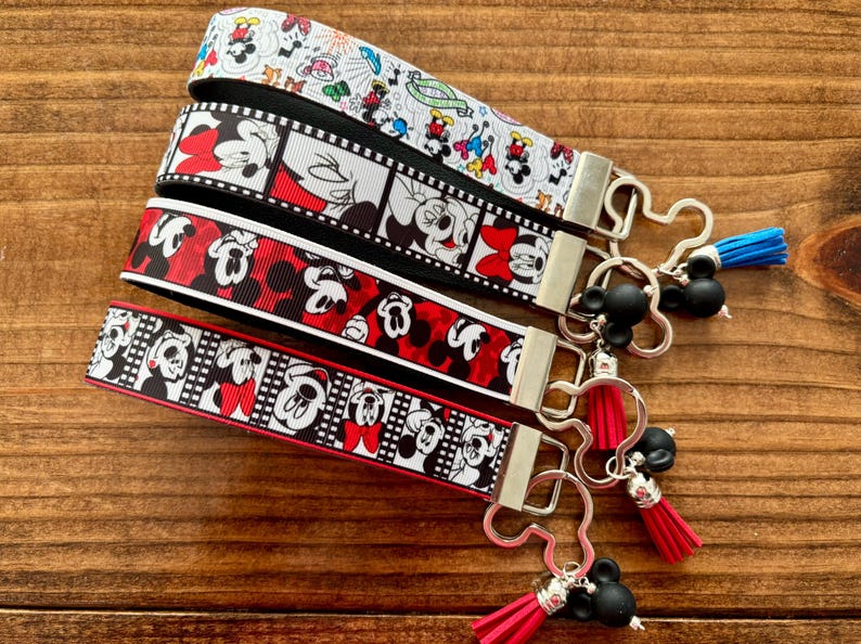 May include: Four key fobs with various Mickey Mouse and Minnie Mouse designs. The key fobs have silver-colored hardware and black Mickey Mouse-shaped charms with tassels. The ribbons feature red, black, and white colors.