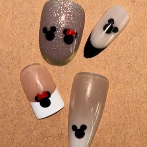 May include: Four false fingernails with a Mickey Mouse and Minnie Mouse design. Two nails have a white tip with a black Mickey Mouse head. One nail has a white tip with a black Mickey Mouse head and a red bow. One nail is a light brown with glitter and has a black Mickey Mouse head and a black Minnie Mouse head with a red bow.