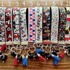 May include: Ten keychains with various designs featuring cartoon characters and Star Wars themes. The keychains have different colored wrist straps with images of Mickey Mouse, Minnie Mouse, and Star Wars characters. Each keychain has a metal clasp and a tassel.